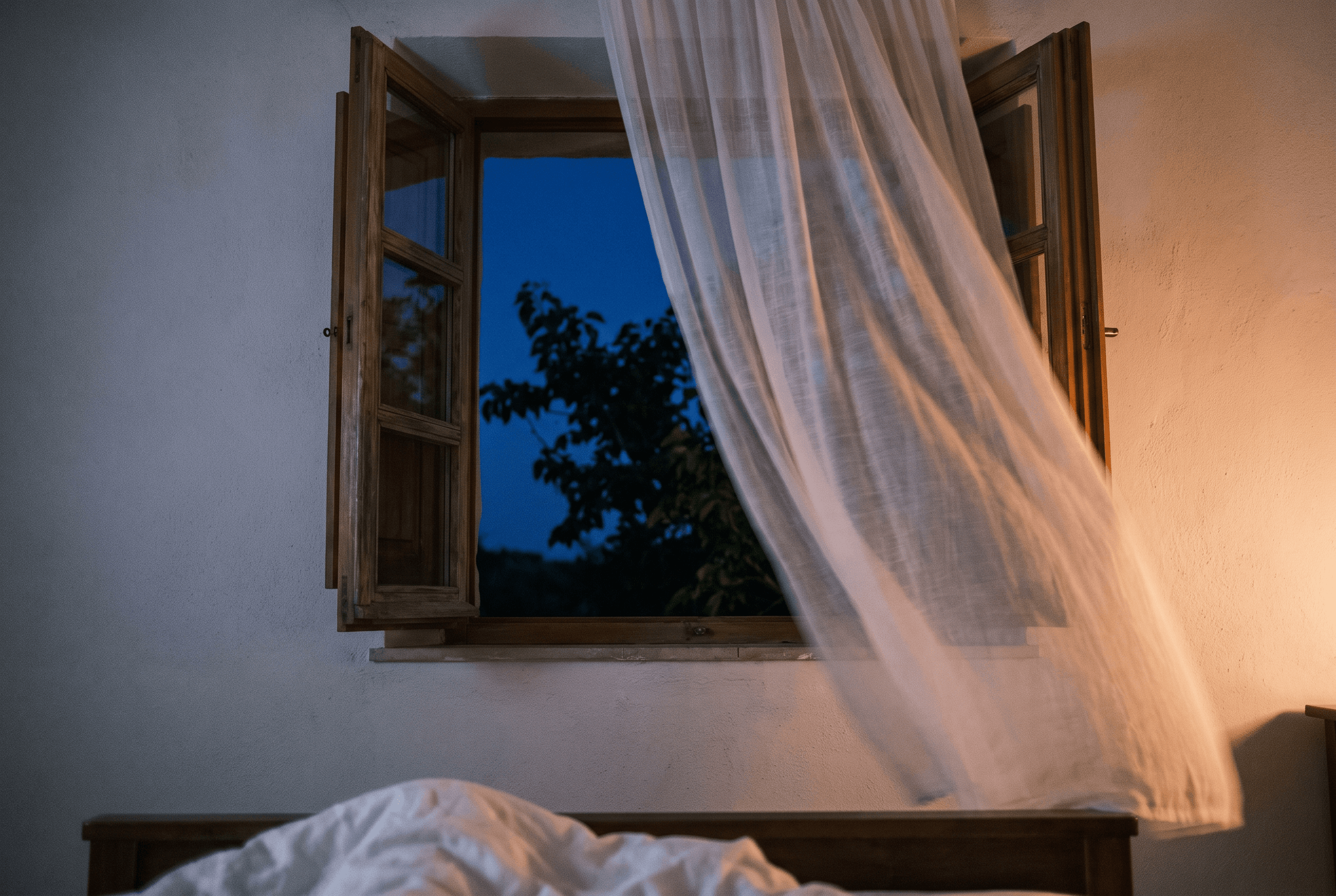 A thin white curtain billowing from an open wooden window at dusk, with rumpled white bedding below.